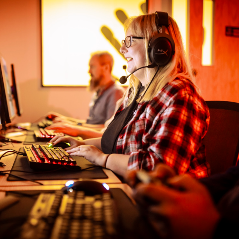 Image shows Jen in a room with red lighting, facing a computer playing a video game. She is wearing a headset with a microphone, like she is playing online with s group of friends. William Cox is in the background, playing on the computer to, but he is slightly faded.